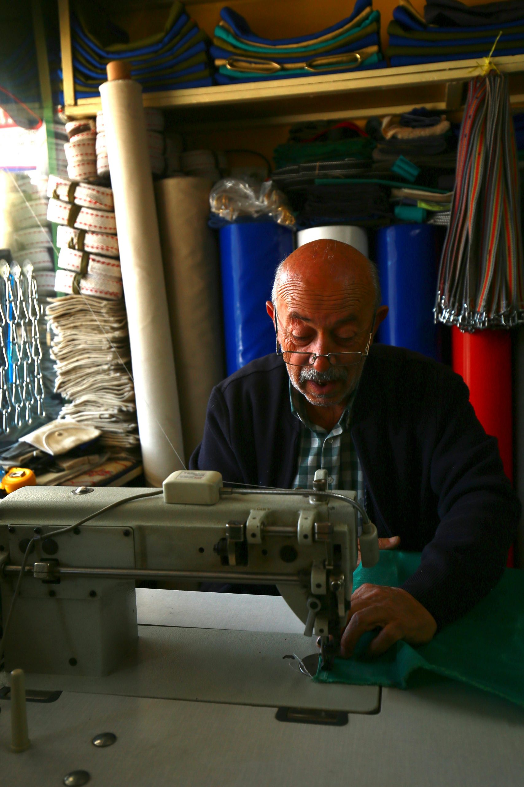 Un homme âgé cousant avec une machine dans un atelier rempli de tissus, mettant en valeur l'artisanat.