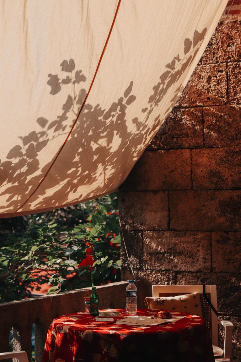 Relaxing outdoor patio with a sunlit fabric canopy casting leaf shadows, perfect for summer afternoons. Patio extérieur relaxant avec un auvent en tissu ensoleillé projetant des ombres de feuilles, parfait pour les après-midis d'été.