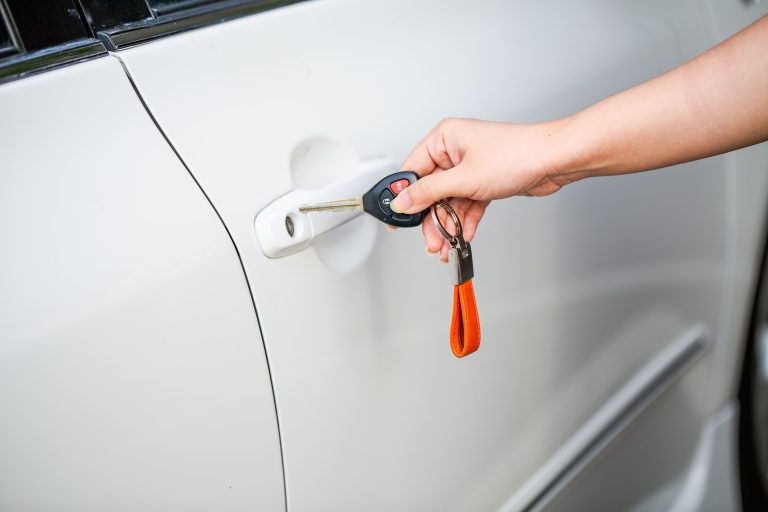 Close-up of a hand unlocking a white car door with a metal key and key fob. Gros plan d'une main déverrouillant une portière de voiture blanche avec une clé métallique et un porte-clés.