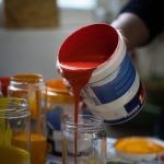 Close-up of a hand pouring vibrant red paint from a bucket into a glass jar, surrounded by other colored paints. Gros plan d'une main versant de la peinture rouge vive d'un seau dans un bocal en verre, entouré d'autres peintures de couleur.