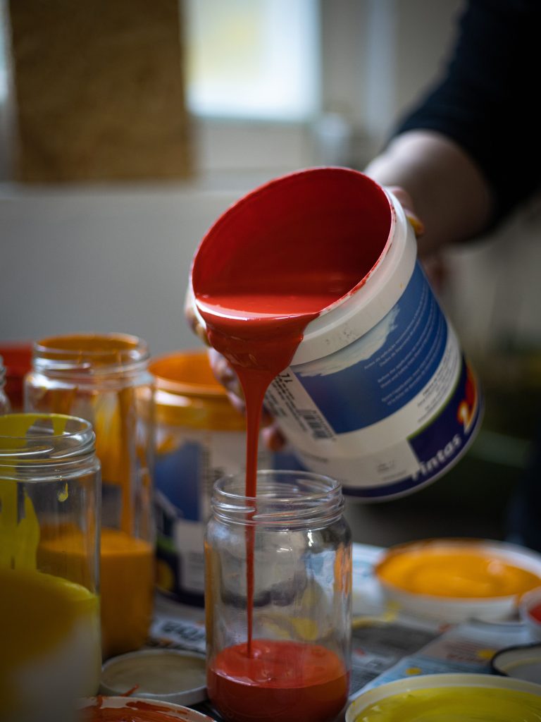 Close-up of a hand pouring vibrant red paint from a bucket into a glass jar, surrounded by other colored paints. Gros plan d'une main versant de la peinture rouge vive d'un seau dans un bocal en verre, entouré d'autres peintures de couleur.